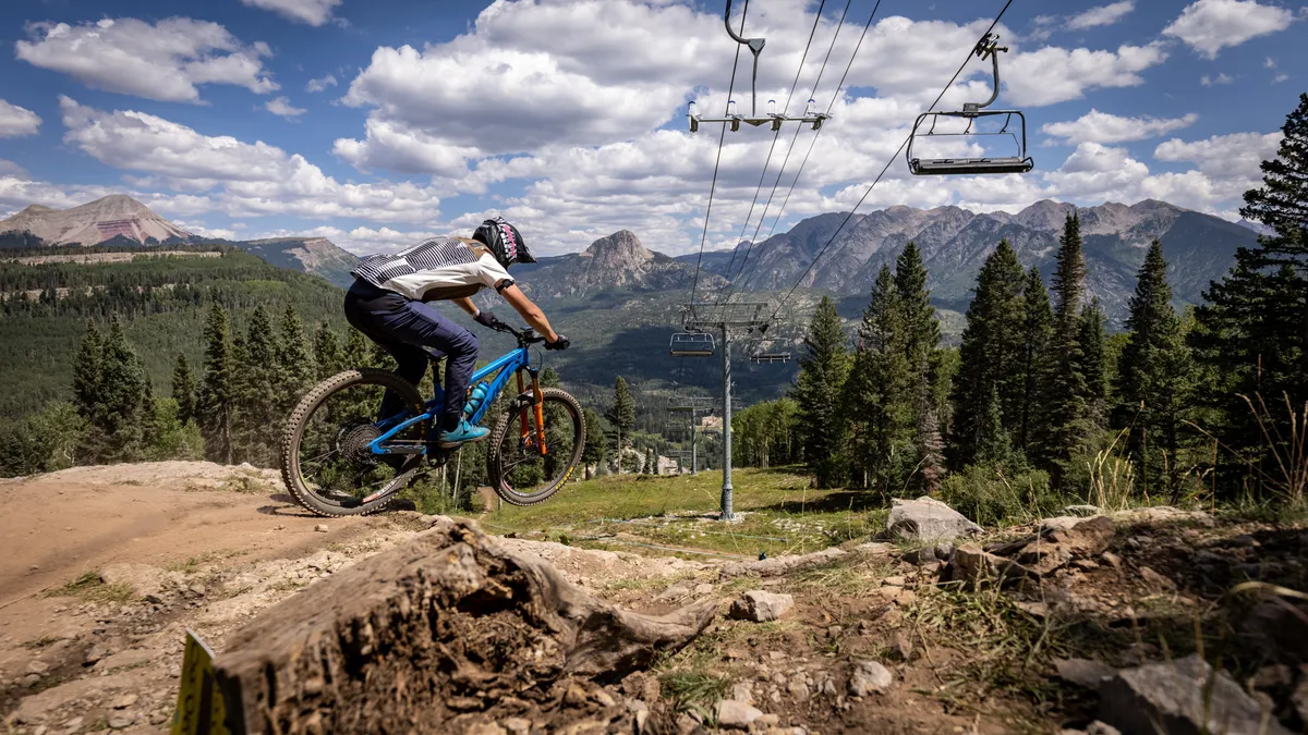 A dynamic exterior shot featuring a mountain biker navigating a trail with an expansive mountain range and a ski lift in the distance. The scene captures summer adventure, with green slopes and clear skies suggesting outdoor activity and exploration.