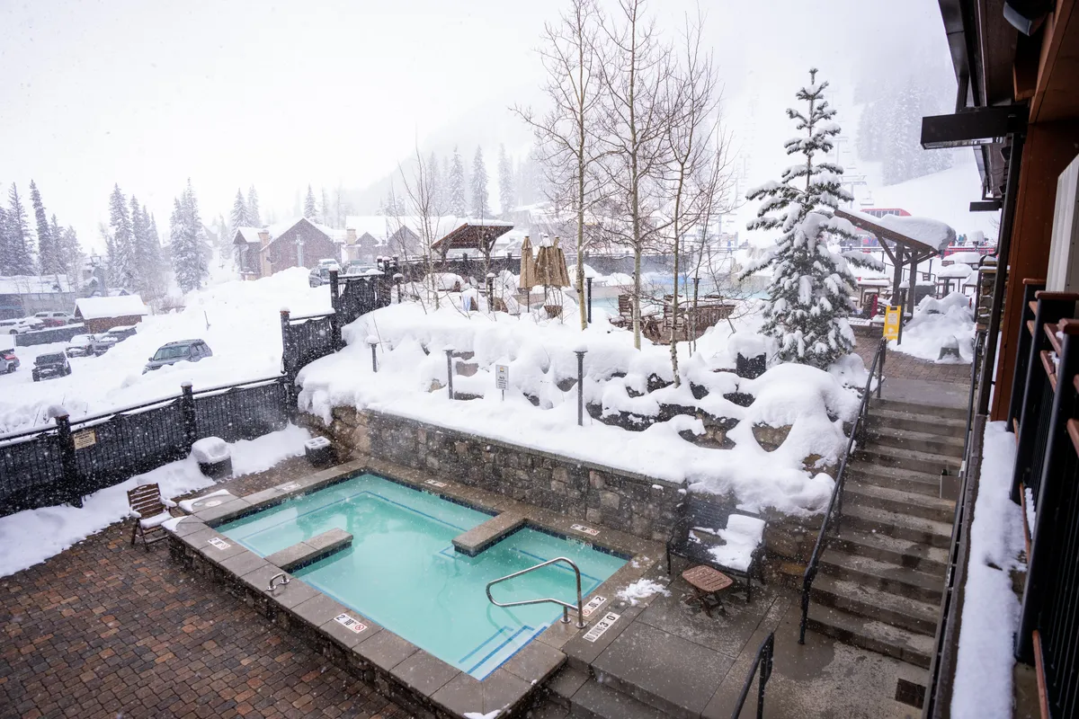 A cozy, snow-covered backyard scene featuring a steaming hot tub set against a panoramic mountain view. Surrounding pine trees and a nearby fire pit complete the tranquil, après-ski atmosphere.