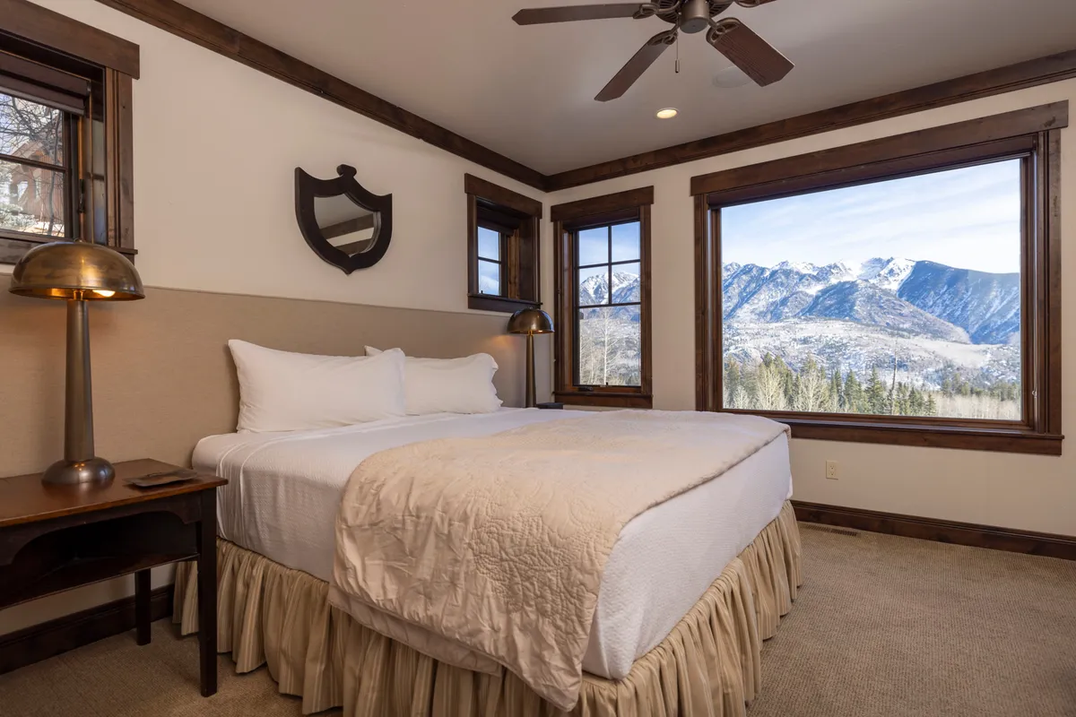 A serene bedroom with a bed facing large windows, allowing natural light and scenic mountain vistas to fill the space. The inclusion of a table lamp and simple décor emphasizes calm and restful nights.