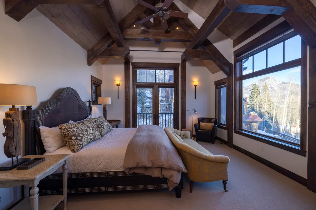 The master suite with a king-sized bed positioned to overlook large windows showcasing mountain views. The room features elegant wooden ceiling beams and a cozy armchair, enhancing the tranquil, private atmosphere.