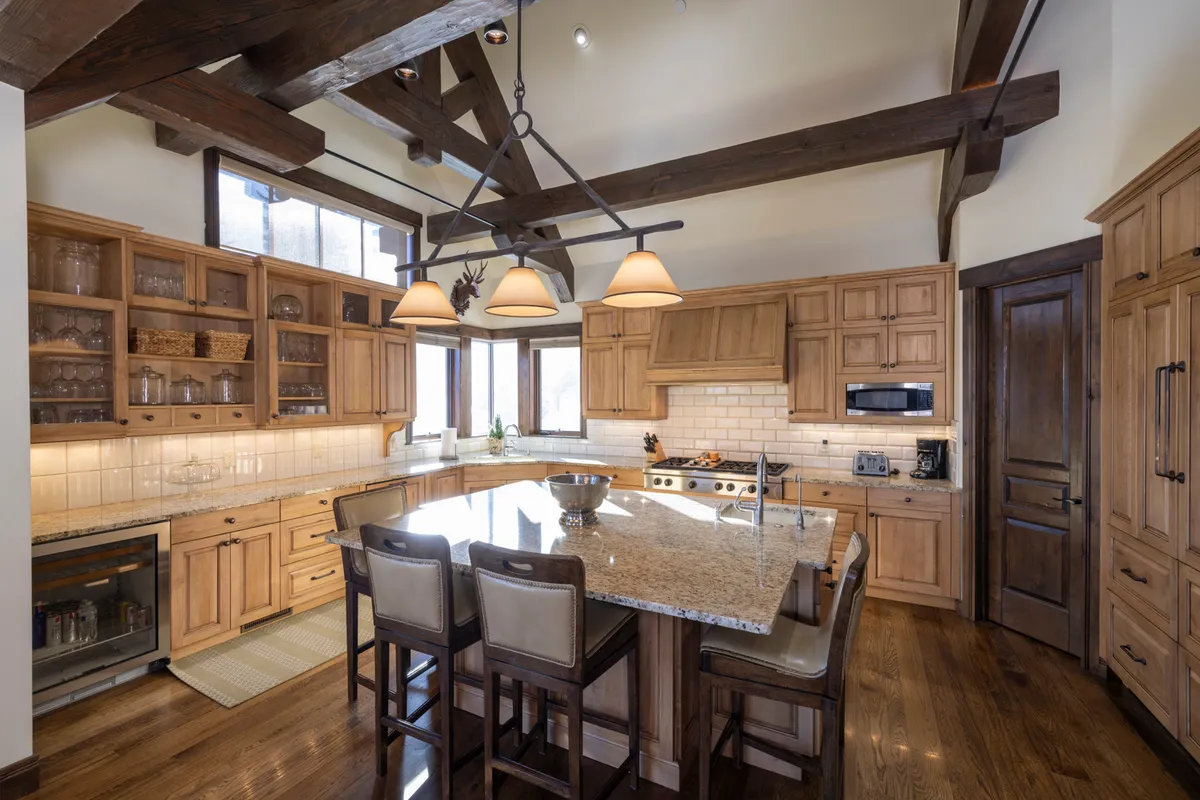 This image showcases a spacious kitchen with a large central island, wooden cabinetry, exposed beams, and abundant natural light streaming through the windows. The kitchen is well-appointed, featuring ample workspace and a welcoming layout ideal for culinary activities and social gatherings.