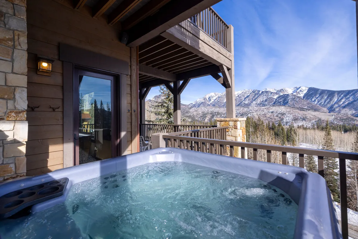 This image showcases a private hot tub perched on a wooden deck, with glass doors leading inside and a breathtaking mountain view in the background. The snowy landscape and tranquil atmosphere evoke a sense of elevated relaxation and immersion in nature.