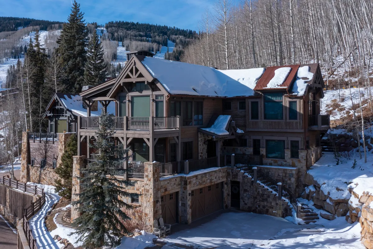 The image showcases a grand multi-level townhome with a striking wooden balcony and a beautiful stone façade, set against a backdrop of snow-covered ground and tall evergreen trees. The scene is tranquil, highlighting the property’s private decks and inviting exterior, with hints of the winter wonderland that surrounds it.
