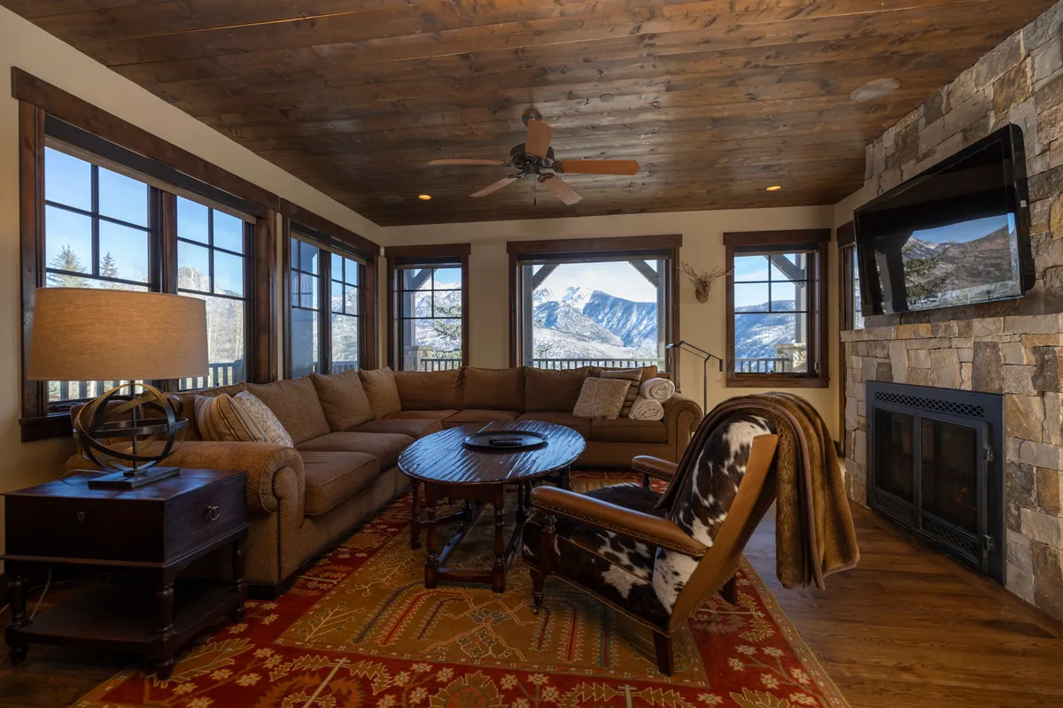 This image features a spacious living room with a large sectional sofa arranged around a stone fireplace. Expansive windows reveal breathtaking mountain views, while a wooden ceiling and decorative rug add warmth and texture. The space is clearly designed for both comfort and gatherings, with ample seating for groups and a cozy ambiance for quieter moments.