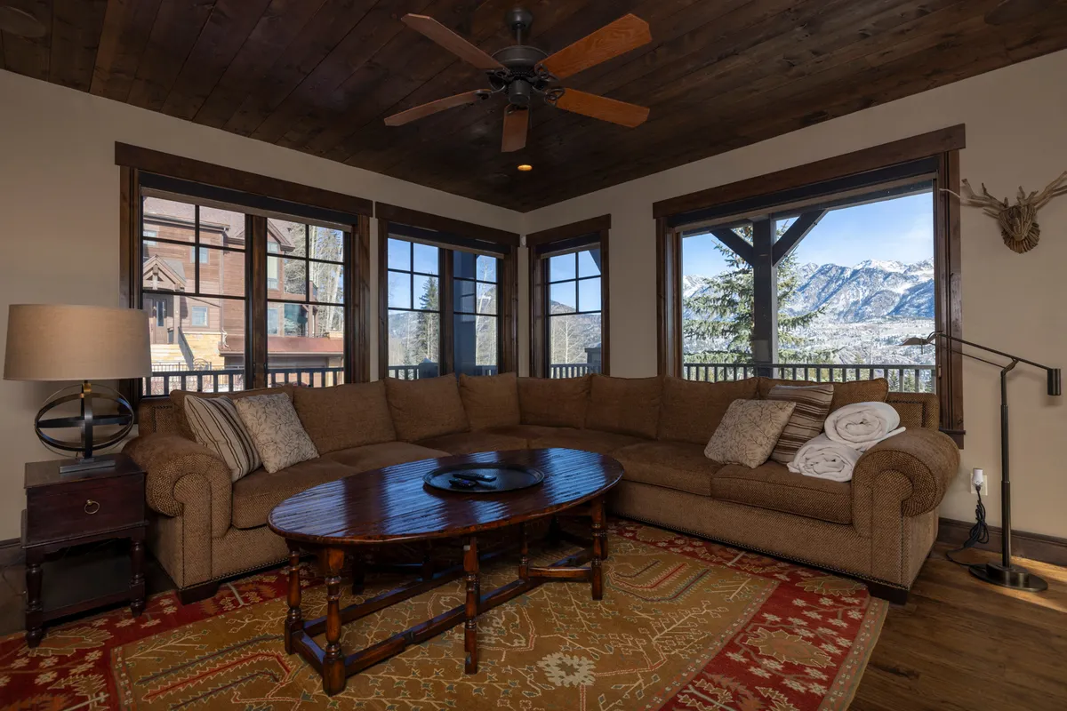 A warm, inviting living space with a large sectional sofa, rustic wooden coffee table, decorative lamp, and windows framing scenic mountain views. The layout suggests a comfortable area for family relaxation or socializing, with plenty of natural light enhancing the cozy atmosphere.