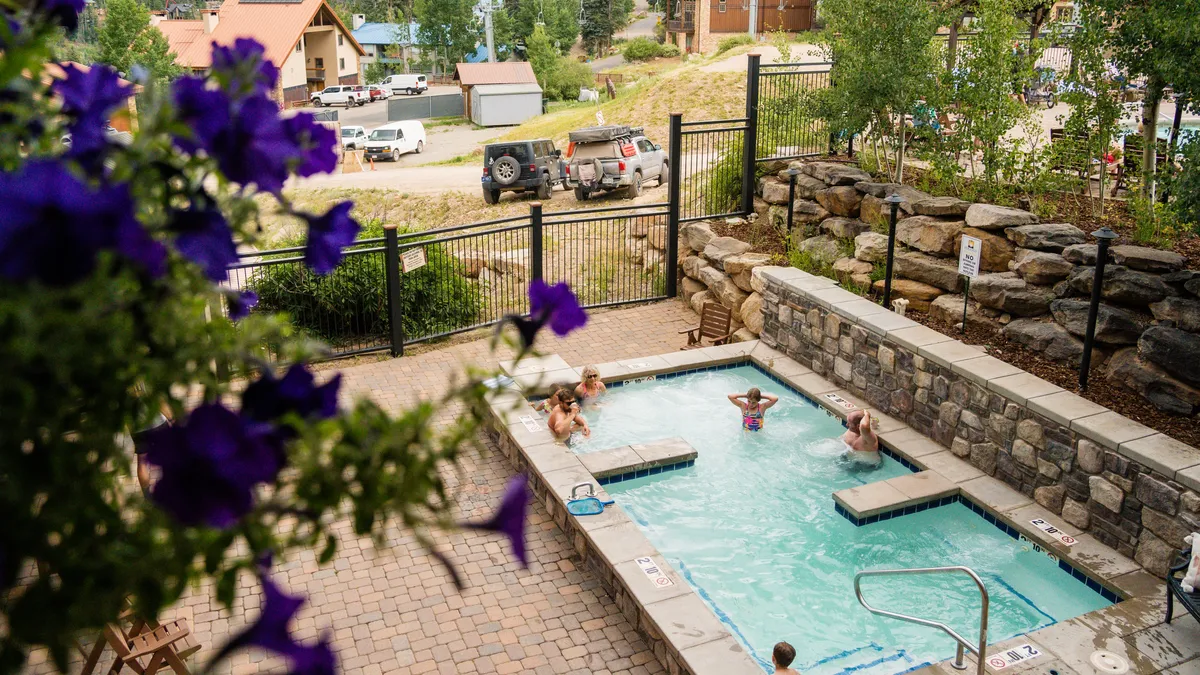 An exterior leisure space featuring a hot tub and swimming pool, surrounded by flowering plants, stone walls, and a fenced area. The setting is lush and inviting, ideal for both lively group activities and relaxing soaks under the open sky, with sunset views enhancing the ambiance.