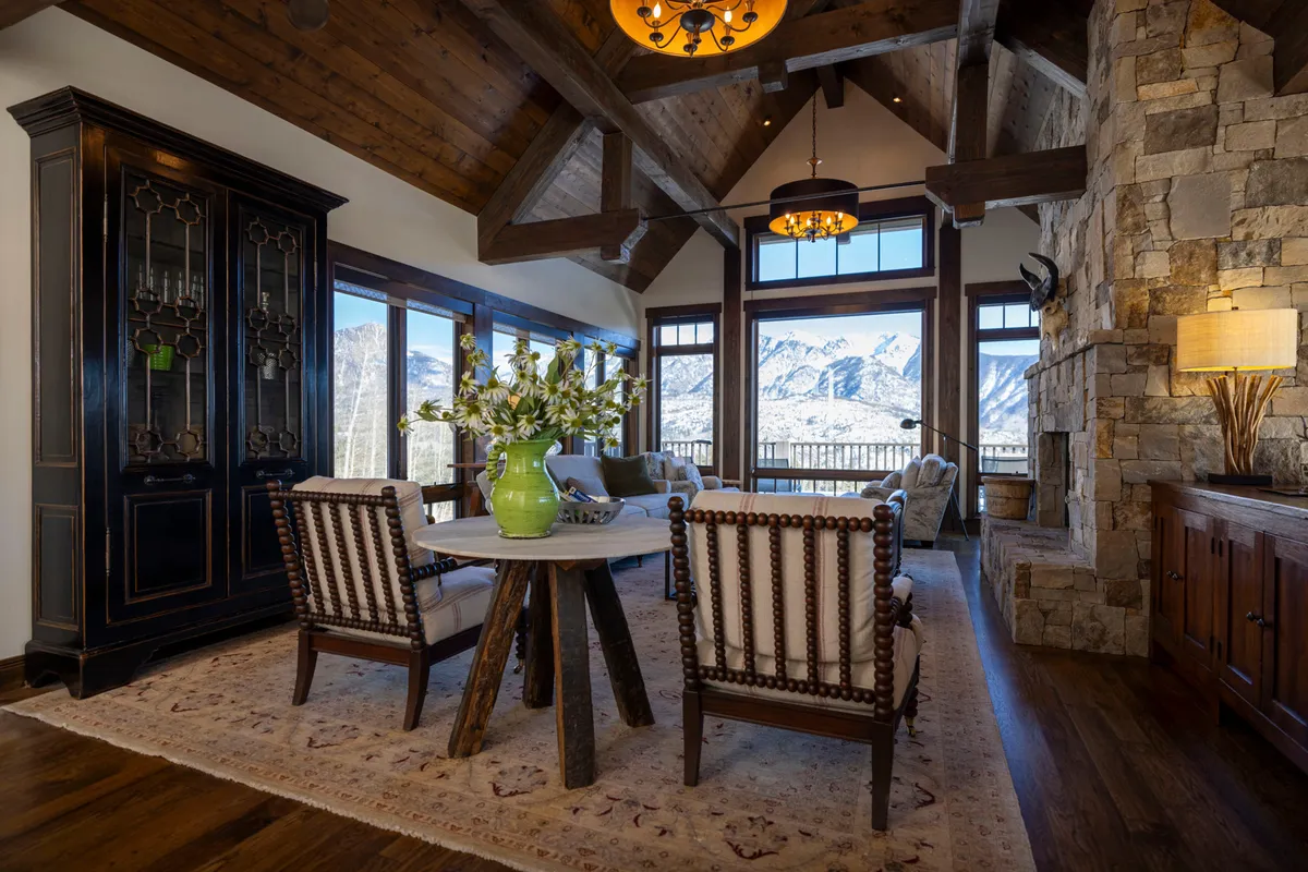 An inviting interior dining and living space, with large windows framing mountain views, exposed wooden beams, a stone fireplace, and a round dining table adorned with flowers. The room is filled with natural light and arranged for group dining, conversation, and relaxation, making it ideal for celebrations and shared meals.