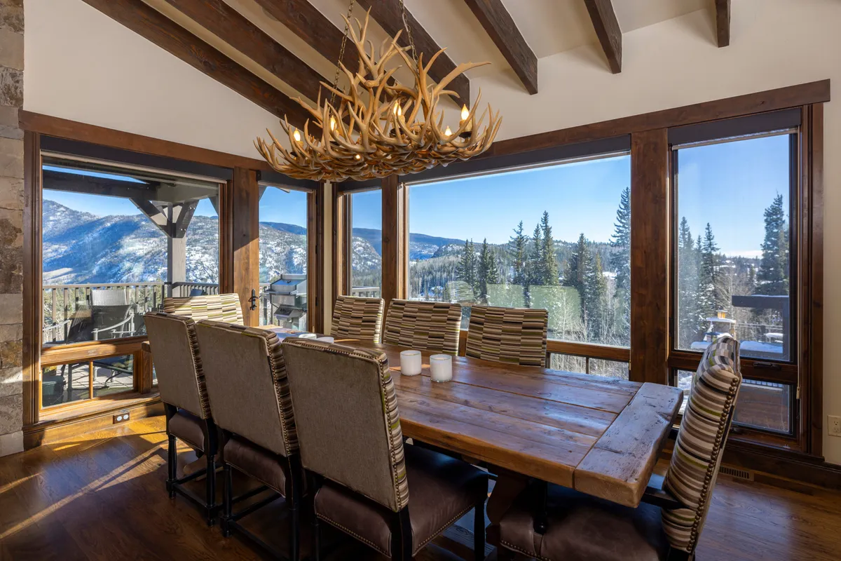This inviting dining area features a large table beneath an antler chandelier, set against expansive windows with a stunning mountain view. The space radiates warmth and is clearly designed for family gatherings or festive celebrations.
