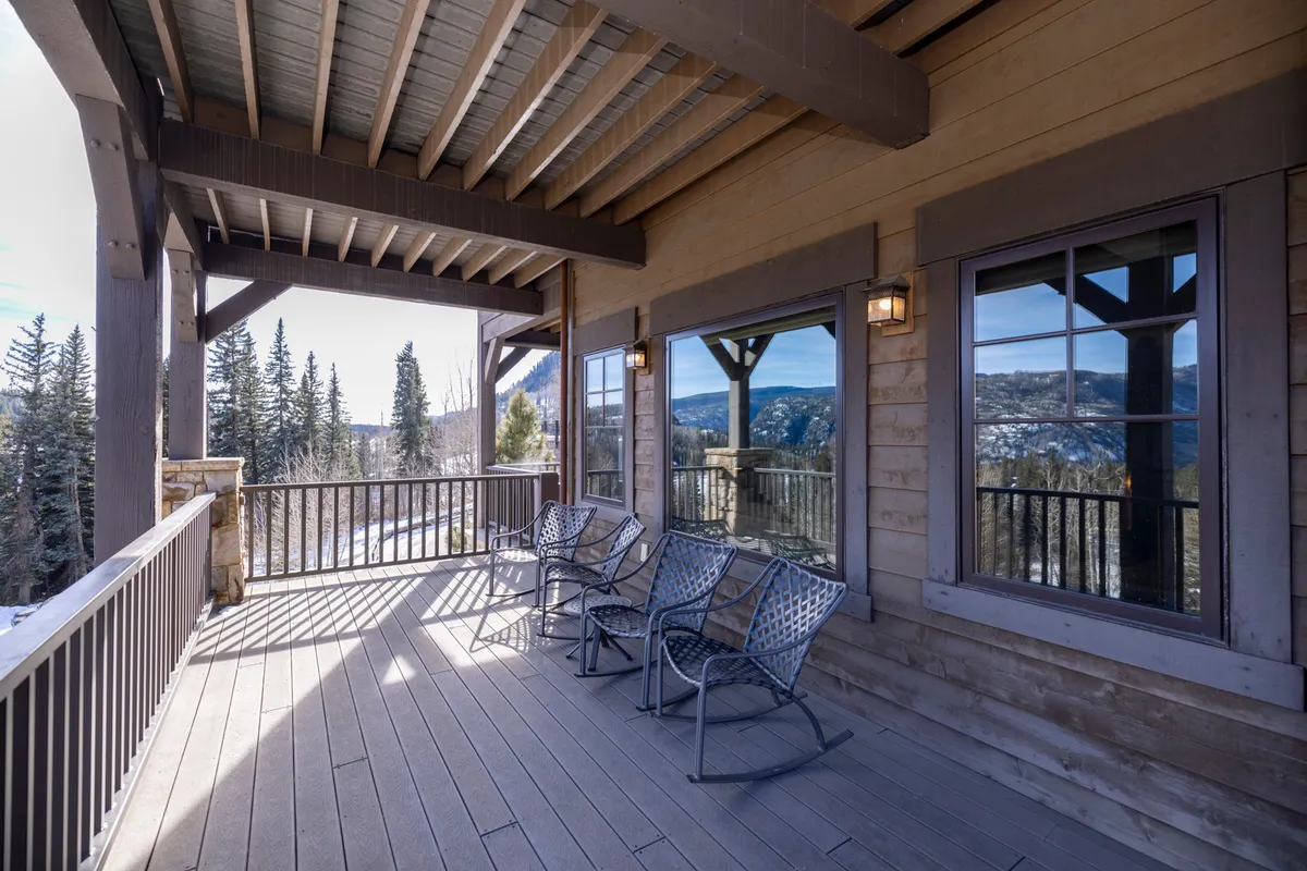 A welcoming exterior scene features wooden rocking chairs on a spacious deck, set against a panoramic mountain view. Large windows reflect the sky and natural surroundings, and the cozy seating arrangement invites relaxation and conversation. The deck is bordered by a wooden railing, with ample space for group gatherings or quiet moments overlooking the landscape.