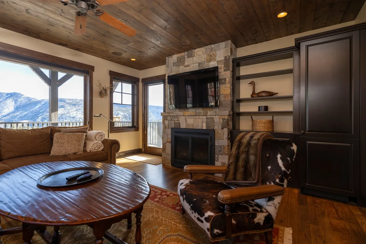 This interior scene features a stone fireplace as its centerpiece, large windows offering stunning mountain views, a rustic wooden coffee table, and a cowhide accent chair. The setup invites both lively gatherings and tranquil moments by the fire while connecting guests to the natural beauty outside.